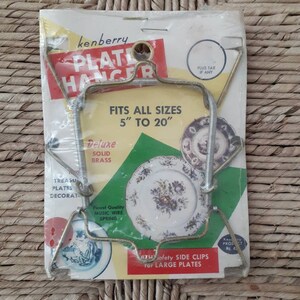 May include: A gold-tone metal plate hanger with a spring mechanism. The packaging says "Kenberry Plate Hanger" and "Fits all sizes 5" to 20". It also says "Deluxe Solid Brass" and "Finest Quality Music Wire Spring".