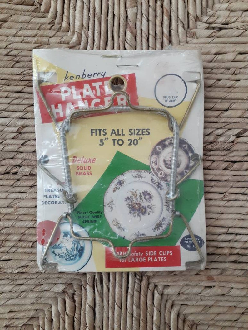 May include: A gold-tone metal plate hanger with a spring mechanism. The packaging says "Kenberry Plate Hanger" and "Fits all sizes 5" to 20". It also says "Deluxe Solid Brass" and "Finest Quality Music Wire Spring".