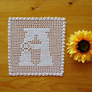 May include: White crocheted doily with a letter "R" design on a wooden surface with a yellow sunflower.