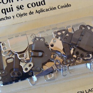 May include: A close-up of a package of sew-on fasteners. The fasteners are silver and black metal and are in a clear plastic package. The package is labeled "Sew-On Fasteners" and "rafe qui se coud" in French. The package also includes the text "e de Gancho y Ojete de Aplicaci&oacute;n Cosido" in Spanish.
