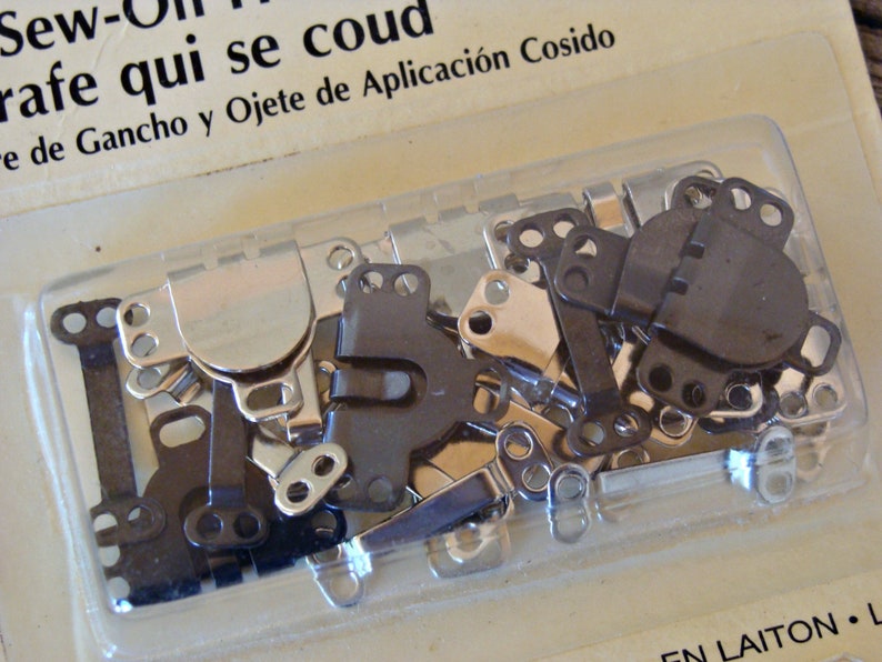 May include: A close-up of a package of sew-on fasteners. The fasteners are silver and black metal and are in a clear plastic package. The package is labeled "Sew-On Fasteners" and "rafe qui se coud" in French. The package also includes the text "e de Gancho y Ojete de Aplicaci&oacute;n Cosido" in Spanish.