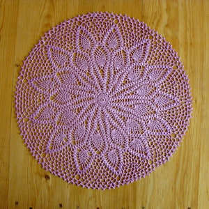 May include: A delicate, circular, lavender crochet doily with a detailed, intricate design. The doily is laid flat on a wooden surface.