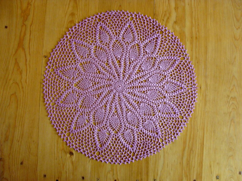 May include: A delicate, circular, lavender crochet doily with a detailed, intricate design. The doily is laid flat on a wooden surface.
