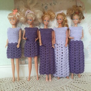 May include: Five dolls wearing purple crocheted dresses. The dresses have a lacy pattern and are all different lengths. The dolls have long blonde hair and are standing on a white surface.