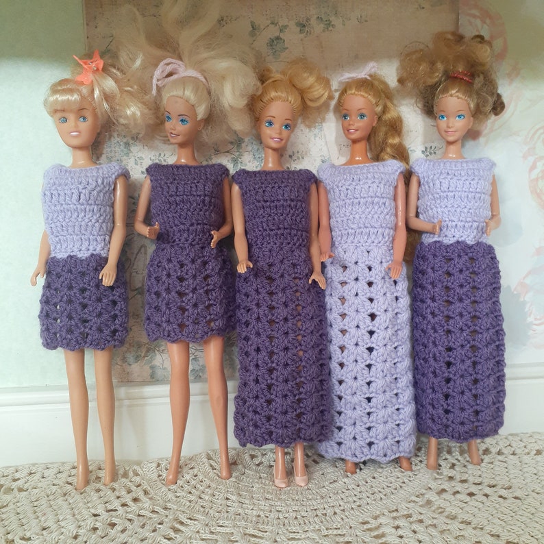 May include: Five dolls wearing purple crocheted dresses. The dresses have a lacy pattern and are all different lengths. The dolls have long blonde hair and are standing on a white surface.