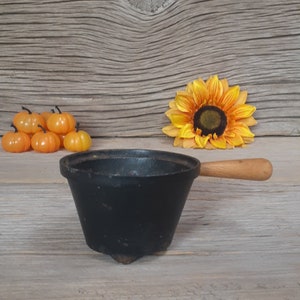 May include: Black cast iron pot with a wooden handle and three legs. The pot is sitting on a wooden surface with small orange pumpkins and a yellow sunflower.