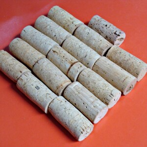 May include: A close-up of a pile of natural cork wine bottle stoppers. The stoppers are arranged in a staggered pattern, with the tops of the stoppers facing the viewer. The stoppers are a light brown color and have a rough, textured surface.