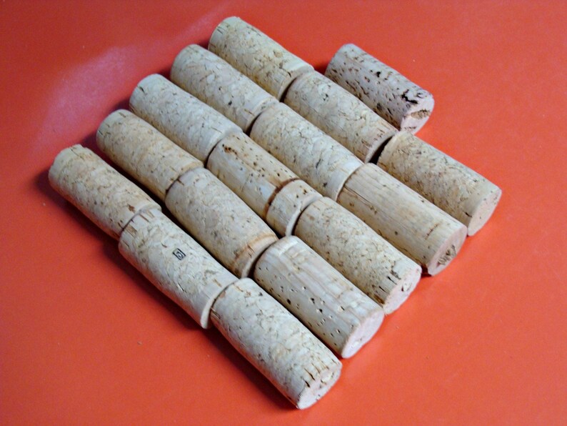 May include: A close-up of a pile of natural cork wine bottle stoppers. The stoppers are arranged in a staggered pattern, with the tops of the stoppers facing the viewer. The stoppers are a light brown color and have a rough, textured surface.