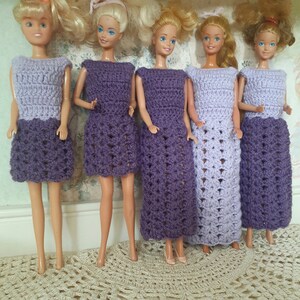 May include: Five Barbie dolls wearing crocheted dresses in shades of purple and lavender. The dresses are all different styles, with some having a more fitted bodice and others having a looser fit. The dolls are all standing on a white surface.