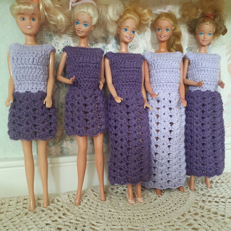 May include: Five Barbie dolls wearing crocheted dresses in shades of purple and lavender. The dresses are all different styles, with some having a more fitted bodice and others having a looser fit. The dolls are all standing on a white surface.