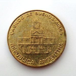 Puede incluir: Una moneda de color dorado con el texto "CABILDO DE BUENOS AIRES REPUBLICA ARGENTINA" alrededor del borde. El centro de la moneda presenta una imagen detallada de un edificio.