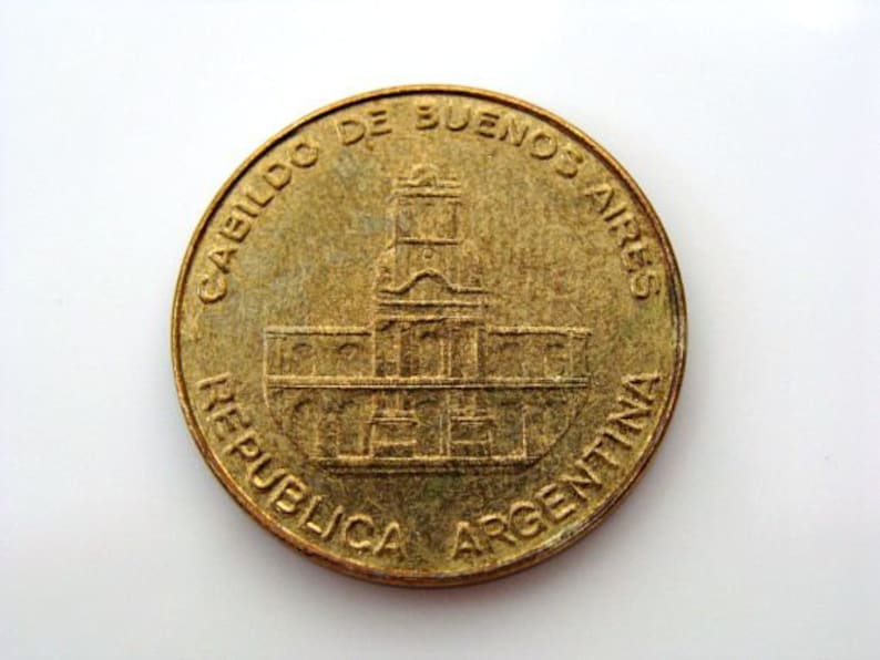 Puede incluir: Una moneda de color dorado con el texto "CABILDO DE BUENOS AIRES REPUBLICA ARGENTINA" alrededor del borde. El centro de la moneda presenta una imagen detallada de un edificio.
