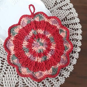 May include: A red, pink, and green crocheted pot holder with a loop handle. The pot holder is circular and has a scalloped edge.