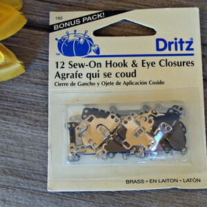 May include: A package of 12 sew-on hook and eye closures in brass. The package is labeled "Dritz" and says "12 Sew-On Hook & Eye Closures" and "Agrafe qui se coud".