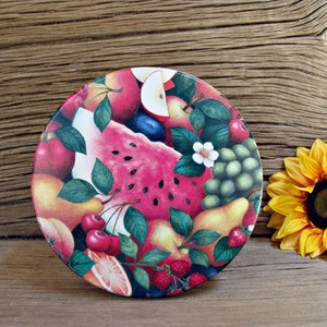 May include: A round ceramic plate with a colorful fruit design. The plate features a watermelon, cherries, grapes, pears, and strawberries. The plate is on a wooden surface.
