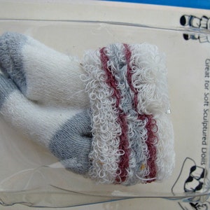 May include: A pair of white doll socks with gray and burgundy stripes. The socks are made of a soft, fuzzy material. "Doll Socks" is printed on the packaging. Great for soft sculpted dolls.