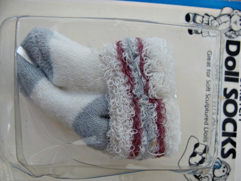May include: A pair of white doll socks with gray and burgundy stripes. The socks are made of a soft, fuzzy material. "Doll Socks" is printed on the packaging. Great for soft sculpted dolls.