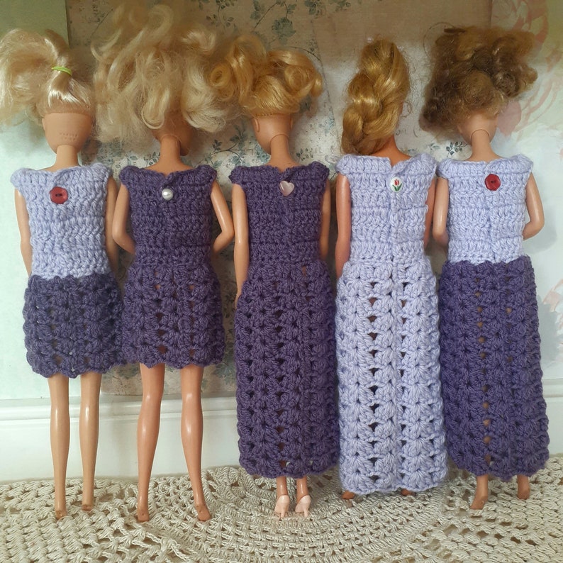 May include: Five dolls wearing crocheted dresses in shades of purple and lavender. The dresses have different lengths and styles, with some featuring a scalloped hemline. The dolls have blonde hair and are standing on a light brown surface.