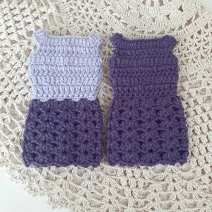 May include: Two crocheted doll dresses, one with a lavender bodice and a purple skirt, and the other entirely purple.