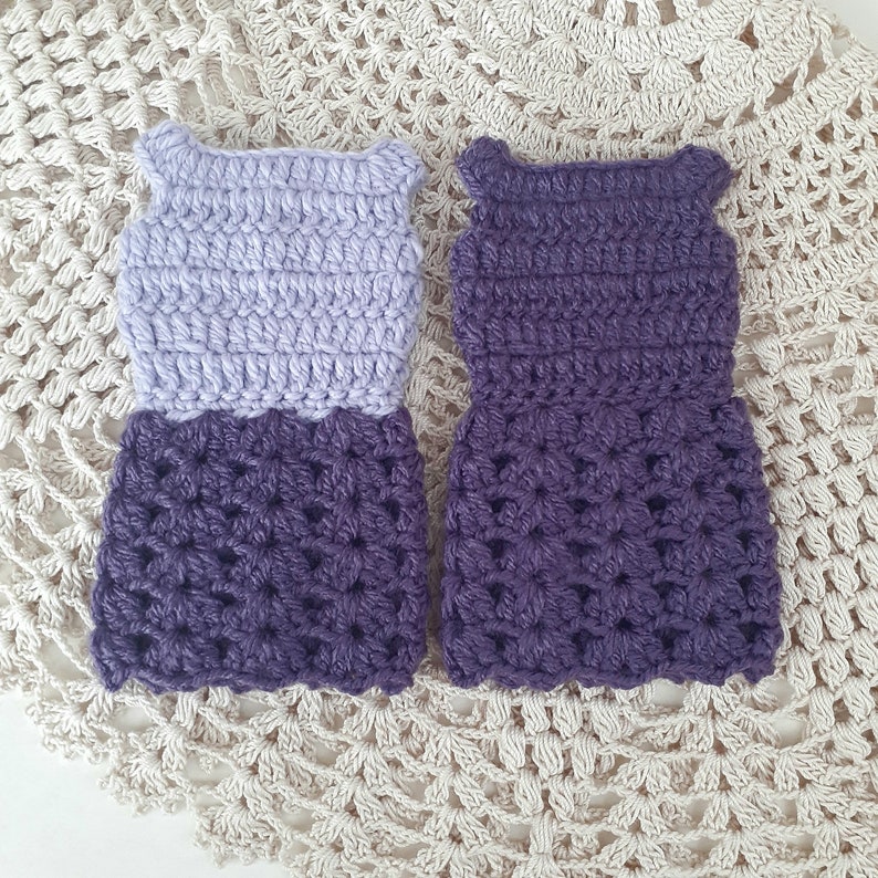 May include: Two crocheted doll dresses, one with a lavender bodice and a purple skirt, and the other entirely purple.