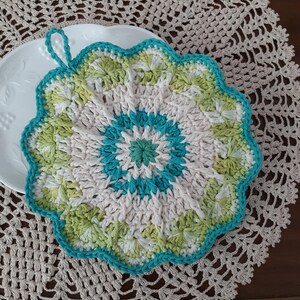 May include: A crocheted pot holder with a scalloped edge in shades of white, green, and turquoise. The pot holder has a loop handle.