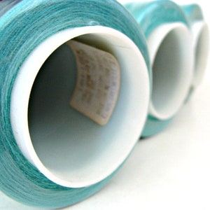 以下が含まれることがあります： 白いプラスチックのボビンに巻き付けられたターコイズブルーの糸の巻き糸のクローズアップ。糸はきつく巻き付けられており、滑らかで均一な質感です。