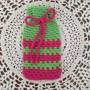 May include: A handmade crochet pouch in green and pink, with a drawstring closure. The pouch features horizontal stripes of green and pink, and a pink drawstring. The pouch is sitting on a white, lacy doily.