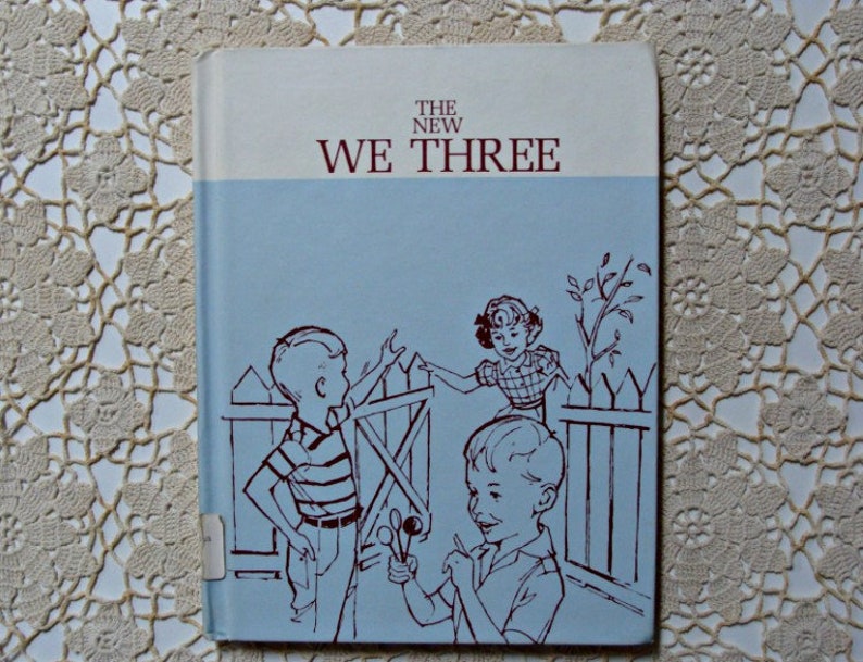 Może przedstawiać: Vintage książka zatytułowana "The New We Three" z jasnoniebieską okładką. Okładka zawiera ilustracje dzieci w pobliżu drewnianego płotu, narysowane brązowym tuszem.