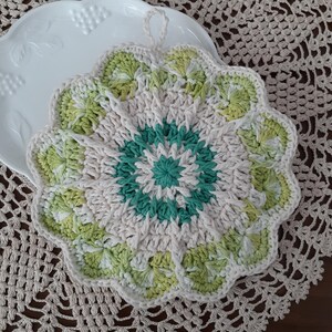 May include: A crocheted white, green, and cream-colored doily with a scalloped edge. The doily has a circular design with a central flower motif.