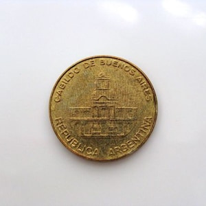 Puede incluir: Una moneda de color dorado con la inscripci&oacute;n "CABILDO DE BUENOS AIRES REPUBLICA ARGENTINA" alrededor del borde. El centro de la moneda presenta una imagen detallada de un edificio.