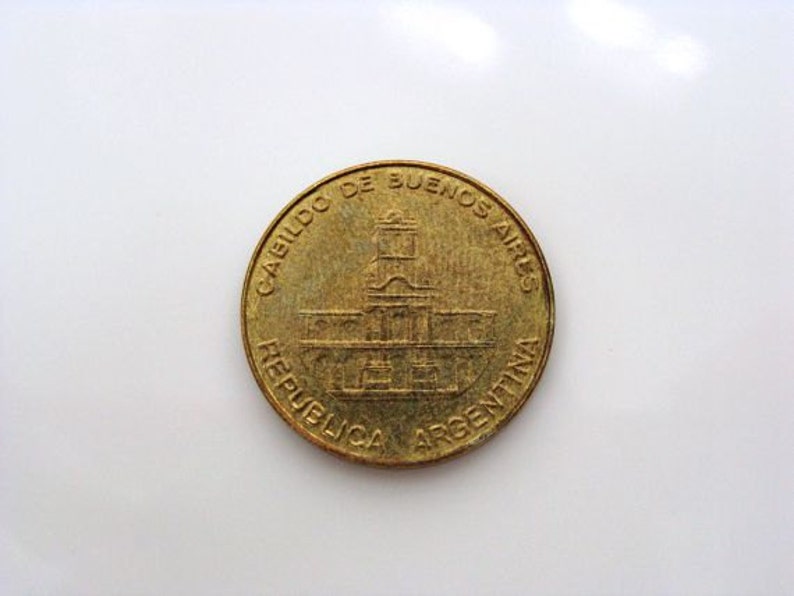 Puede incluir: Una moneda de color dorado con la inscripci&oacute;n "CABILDO DE BUENOS AIRES REPUBLICA ARGENTINA" alrededor del borde. El centro de la moneda presenta una imagen detallada de un edificio.