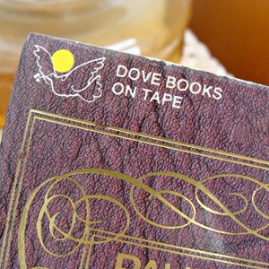 May include: Close-up of a book cover with the text "DOVE BOOKS ON TAPE". The cover is a deep burgundy colour with gold decorative accents and a white dove logo with a yellow circle. The book appears to be vintage.