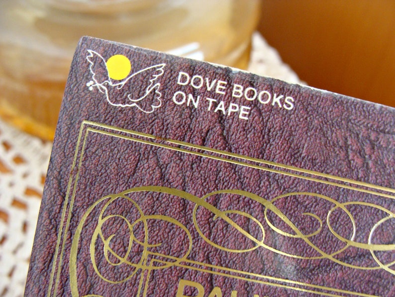 May include: Close-up of a book cover with the text "DOVE BOOKS ON TAPE". The cover is a deep burgundy colour with gold decorative accents and a white dove logo with a yellow circle. The book appears to be vintage.