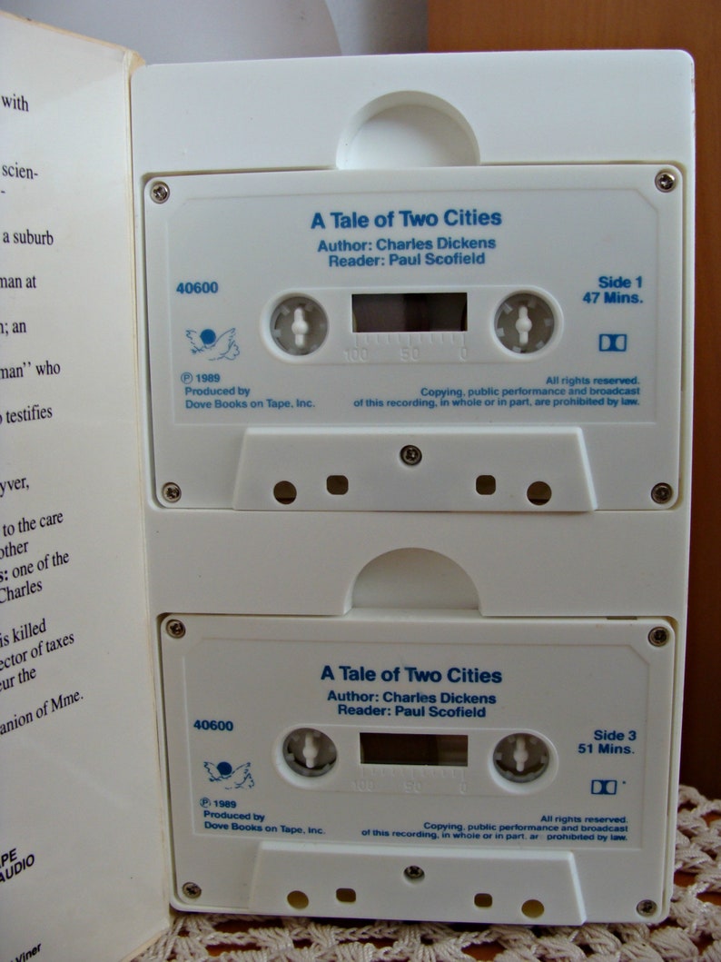 May include: Two white cassette tapes, featuring the title "A Tale of Two Cities" by Charles Dickens. The tapes have blue text and a dove logo, with "Side 1 47 Mins." and "Side 3 51 Mins." printed on them. The tapes are in a white case.