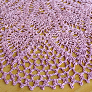 May include: A delicate, crocheted doily in a soft pink hue. The doily features a circular design with intricate lacework and a scalloped edge.