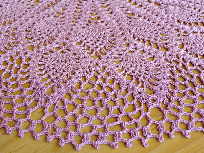 May include: A delicate, crocheted doily in a soft pink hue. The doily features a circular design with intricate lacework and a scalloped edge.