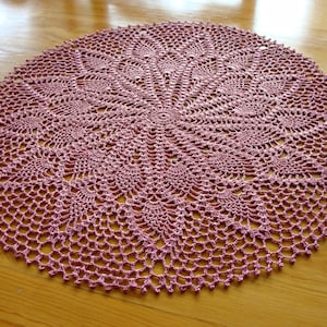 May include: A pink crocheted doily with a delicate floral pattern. The doily is circular and has a lacy design.