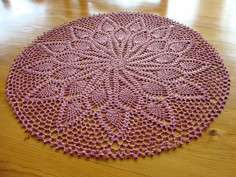 May include: A pink crocheted doily with a delicate floral pattern. The doily is circular and has a lacy design.