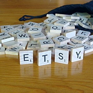 May include: A close-up of a pile of wooden letter tiles, with the word "ETSY" spelled out in the center. The tiles are in a black fabric bag, and the background is a wooden table.
