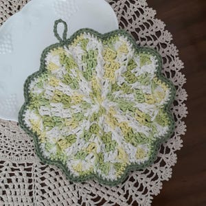 May include: A crocheted pot holder with a scalloped edge in shades of green, white, and yellow. The pot holder has a loop for hanging.
