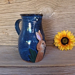 May include: A blue ceramic pitcher with a brown rabbit illustration and green leaves. The pitcher has a handle and is on a wooden surface.