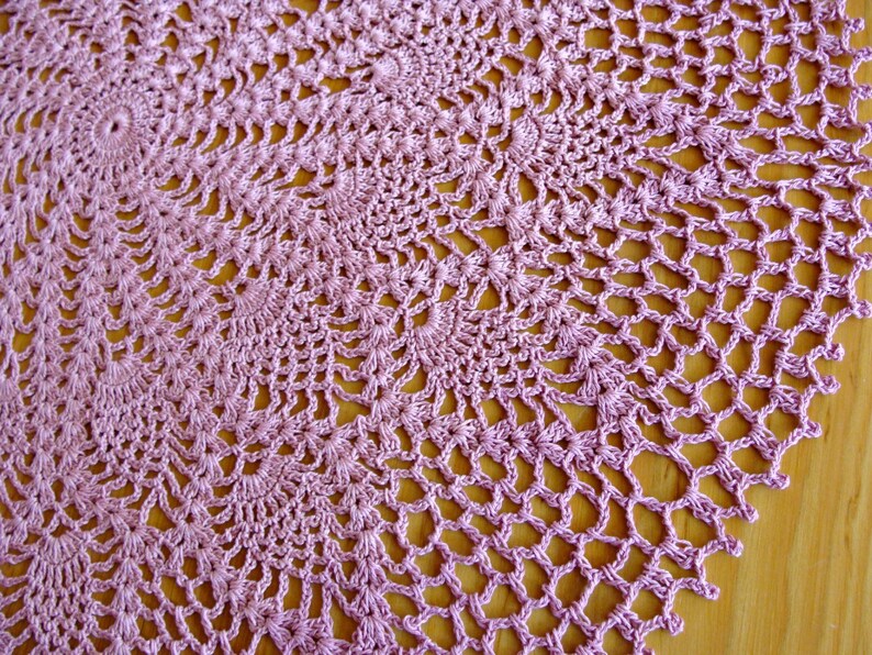 May include: A delicate, round, crocheted doily in a pale lavender color. The doily is made with a fine thread and has a delicate, intricate pattern.