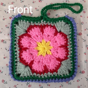 May include: A handmade crochet square with a floral design. The center is yellow, surrounded by pink petals, red accents, and gray leaves. The border is green, blue, and purple, with a green loop for hanging. The word "Front" is written on the top left.