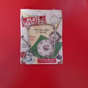 May include: A vintage plate hanger made of solid brass, packaged in a white and green cardboard box. The hanger is designed to hold plates of various sizes, from 5 to 20 inches. The packaging features the brand name "kenberry" and the text "Fits all sizes 5" to 20" Deluxe Solid Brass".