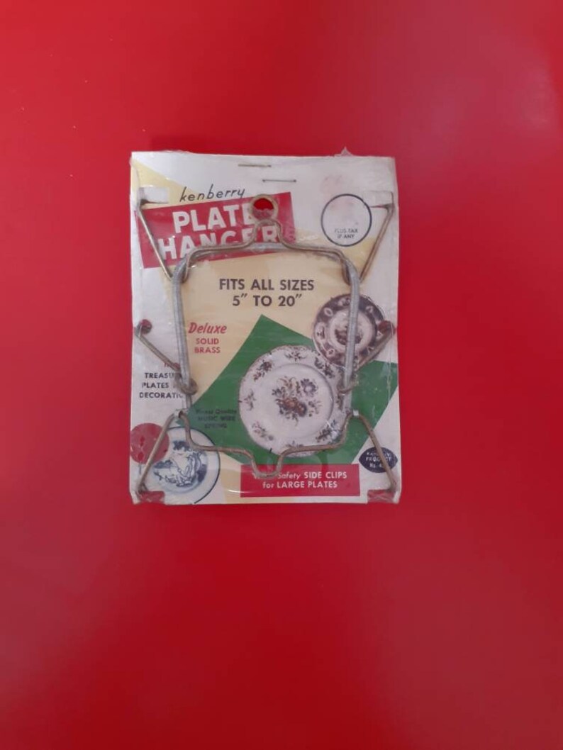 May include: A vintage plate hanger made of solid brass, packaged in a white and green cardboard box. The hanger is designed to hold plates of various sizes, from 5 to 20 inches. The packaging features the brand name "kenberry" and the text "Fits all sizes 5" to 20" Deluxe Solid Brass".