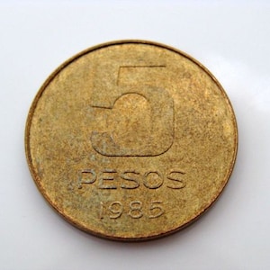 Puede incluir: Una moneda de color dorado con el n&uacute;mero "5" y la palabra "PESOS" inscrita en ella. El a&ntilde;o "1985" tambi&eacute;n es visible en la moneda.