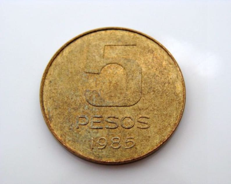 Puede incluir: Una moneda de color dorado con el n&uacute;mero "5" y la palabra "PESOS" inscrita en ella. El a&ntilde;o "1985" tambi&eacute;n es visible en la moneda.