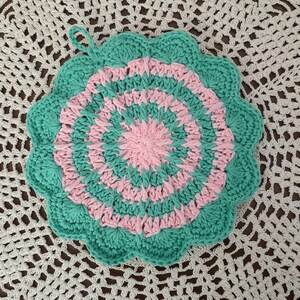 May include: A crocheted pot holder with a pink and green circular design. The pot holder has a scalloped edge and a loop for hanging.