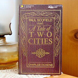 May include: A vintage audio cassette of "A Tale of Two Cities" by Charles Dickens, read by Paul Scofield. The book cover is a deep burgundy with gold lettering and decorative flourishes. The cassette case is visible at the top.