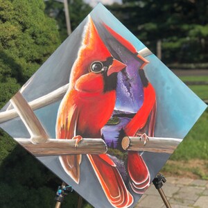 以下が含まれることがあります： 枝に止まっている2羽の赤いカージナルを描いた絵画。カージナルはリアルなスタイルで描かれており、羽毛のディテールや鮮やかな色が表現されています。絵画の背景は濃い青色で、星々の銀河と鳥を通して見える風景が描かれています。